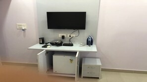 A compact room with a flat-screen TV mounted on the wall, a small refrigerator, a microwave, and a coffee maker on a white desk.