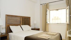 Hotelkamer met een houten hoofdeinde, een bed met wit en bruin beddengoed, een nachtkastje met een lamp, een raam met gordijnen en uitzicht op een gebouw.