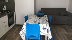 Eine kleine Küche mit Herd, Backofen und Spüle. Ein Esstisch mit blauen Stühlen steht vor einer weißen Wand mit zwei gerahmten Bildern.