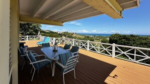 Una terraza de madera con barandilla blanca, una mesa con sillas y vistas al océano y las colinas.