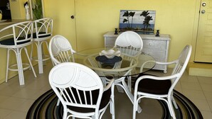 Un comedor con una mesa redonda de vidrio rodeada de sillas de mimbre blanco, un ventilador de techo y una unidad de aire acondicionado en la pared.
