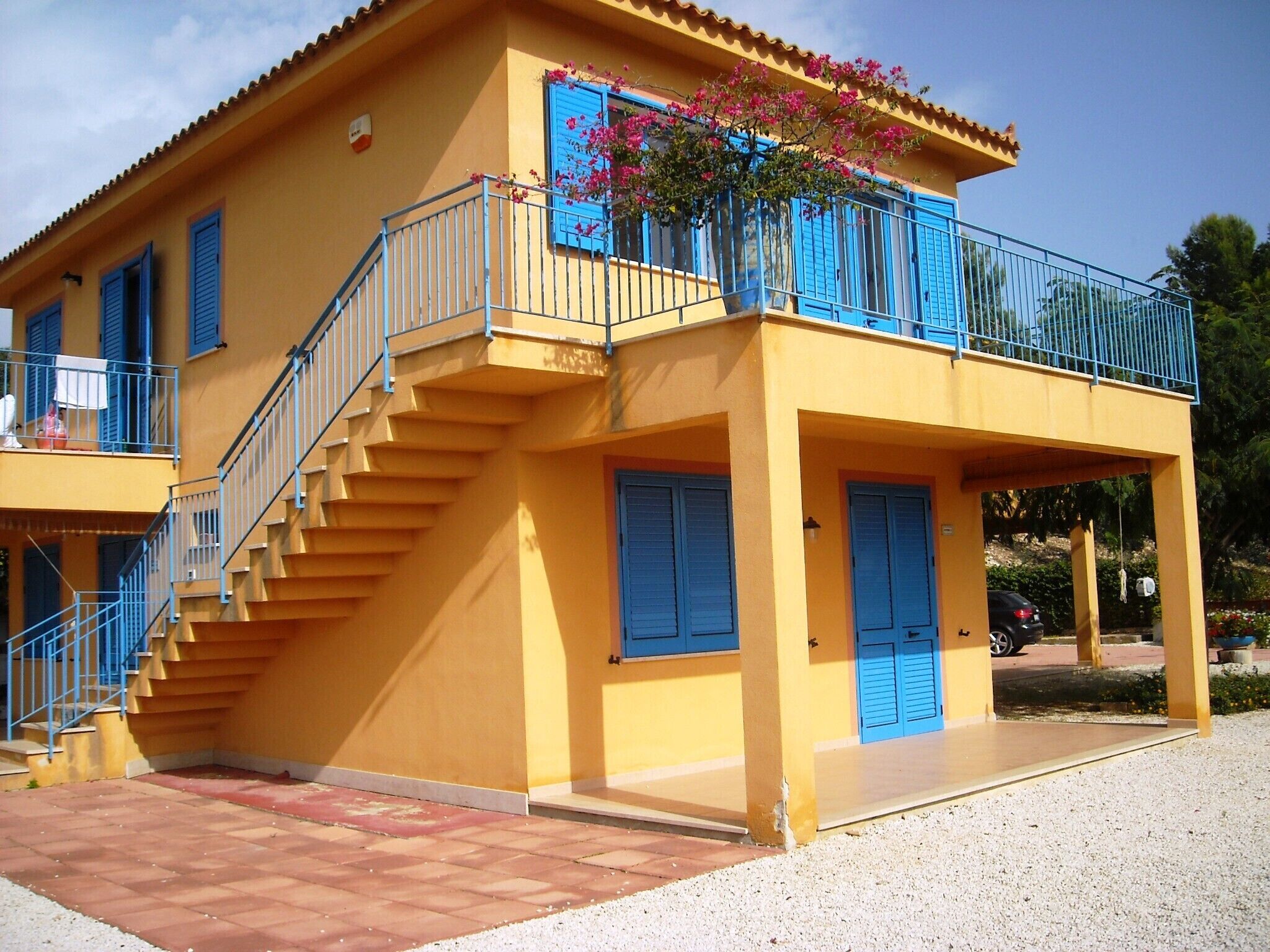 Un edificio amarillo con contraventanas azules, una escalera y un balcón adornado con flores rosadas.