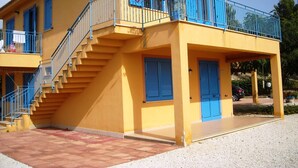 Un edificio amarillo con contraventanas azules, una escalera y un balcón adornado con flores rosadas.