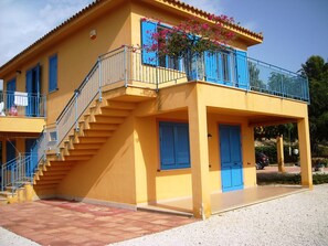 Ein gelbes Gebäude mit blauen Fensterläden, einer Treppe und einem Balkon mit rosa Blumen.