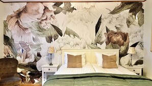 Un dormitorio con un amplio mural floral en la pared, una cama con ropa de cama verde, dos mesitas de noche con lámparas y una silla.