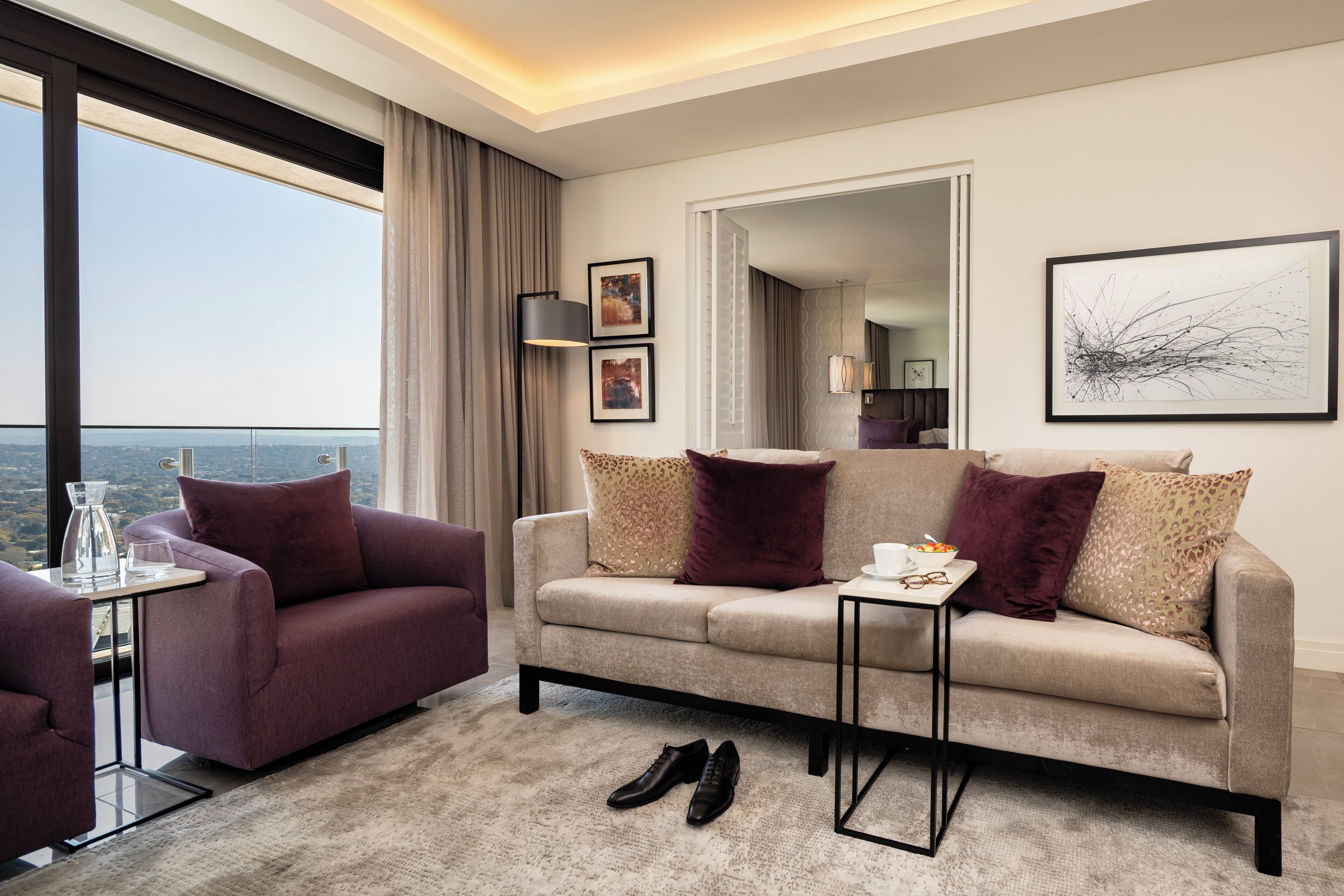 A modern hotel room with a large window, a sofa, a coffee table, and a pair of shoes on the floor.