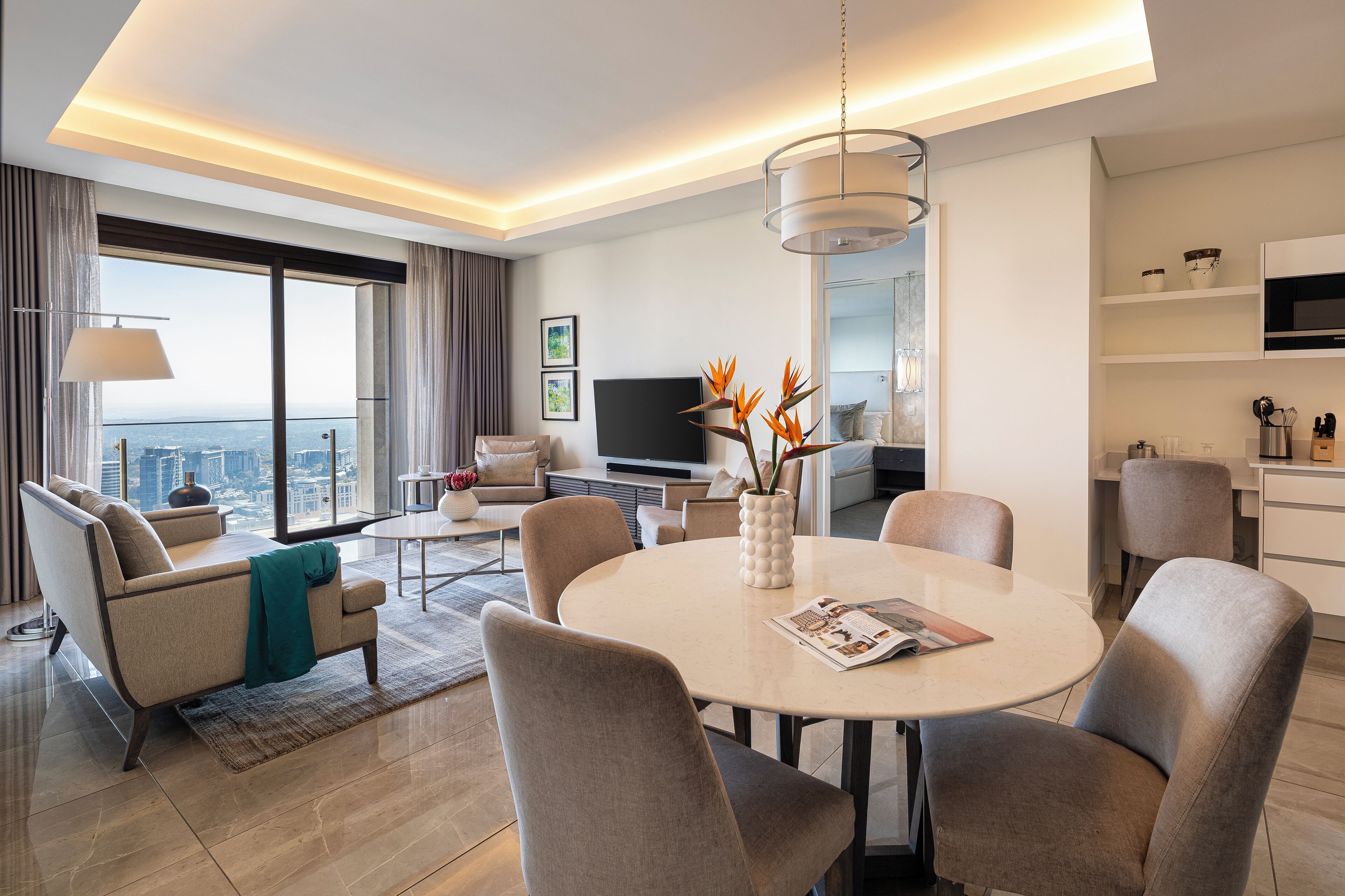 Un salon moderne avec une table à manger ronde, des chaises recouvertes d’un tissu gris, un téléviseur à écran plat et une vue sur la ville.