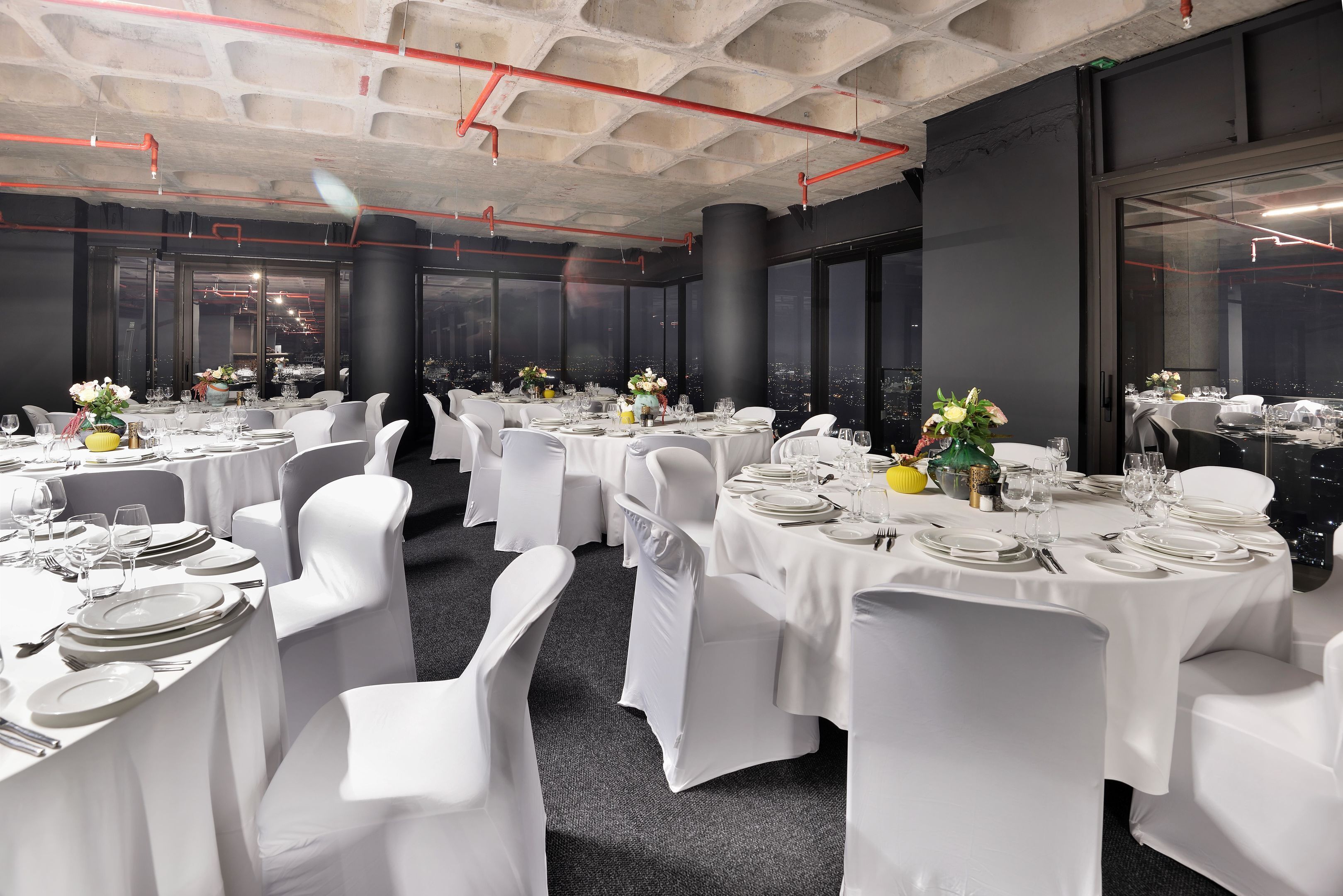 A banquet hall with multiple round tables set for dining, white chairs, and large windows offering a city view.