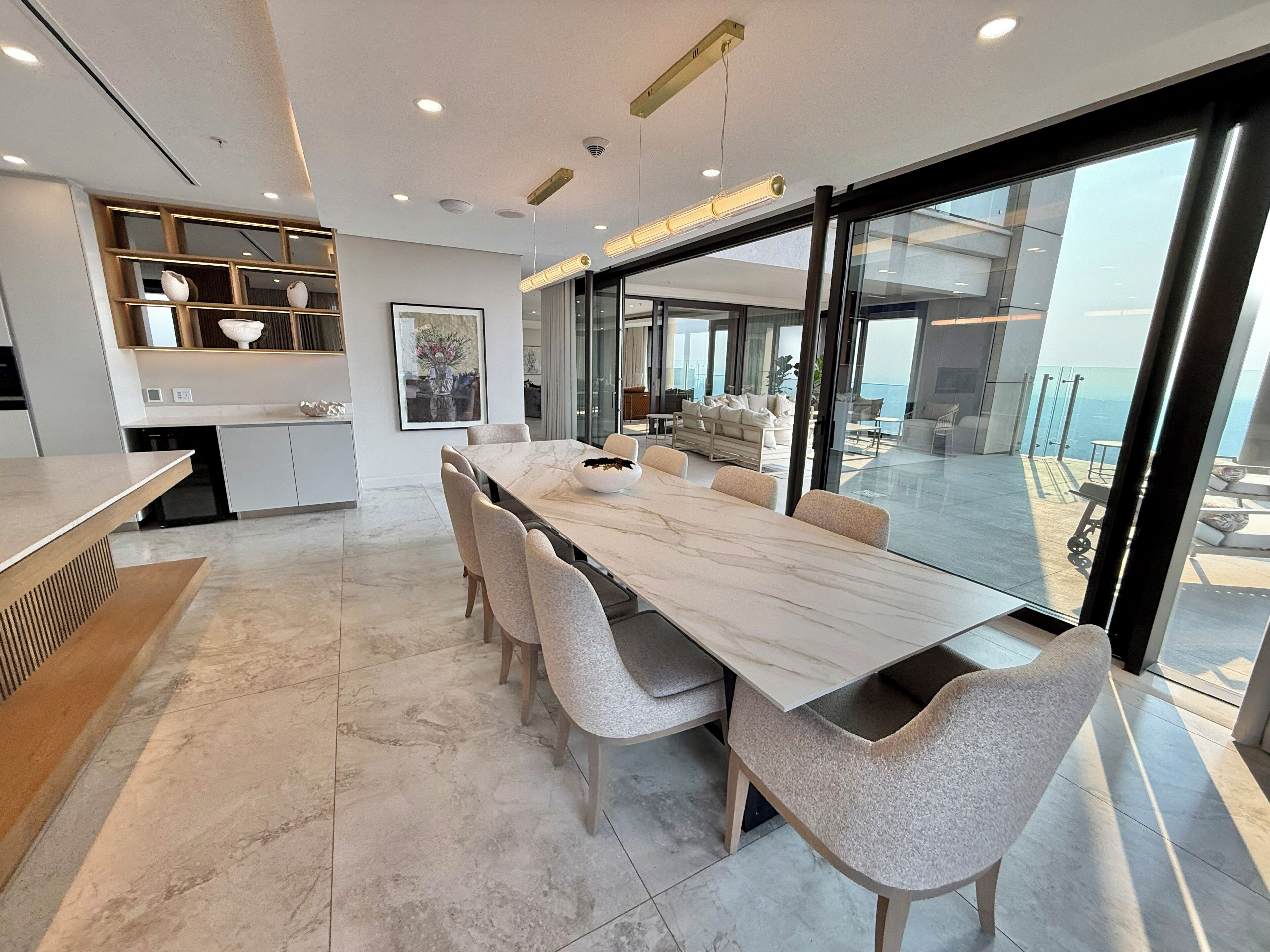 A modern kitchen with a marble countertop, a dining area with a long table and chairs, and a view of the ocean through large glass doors.