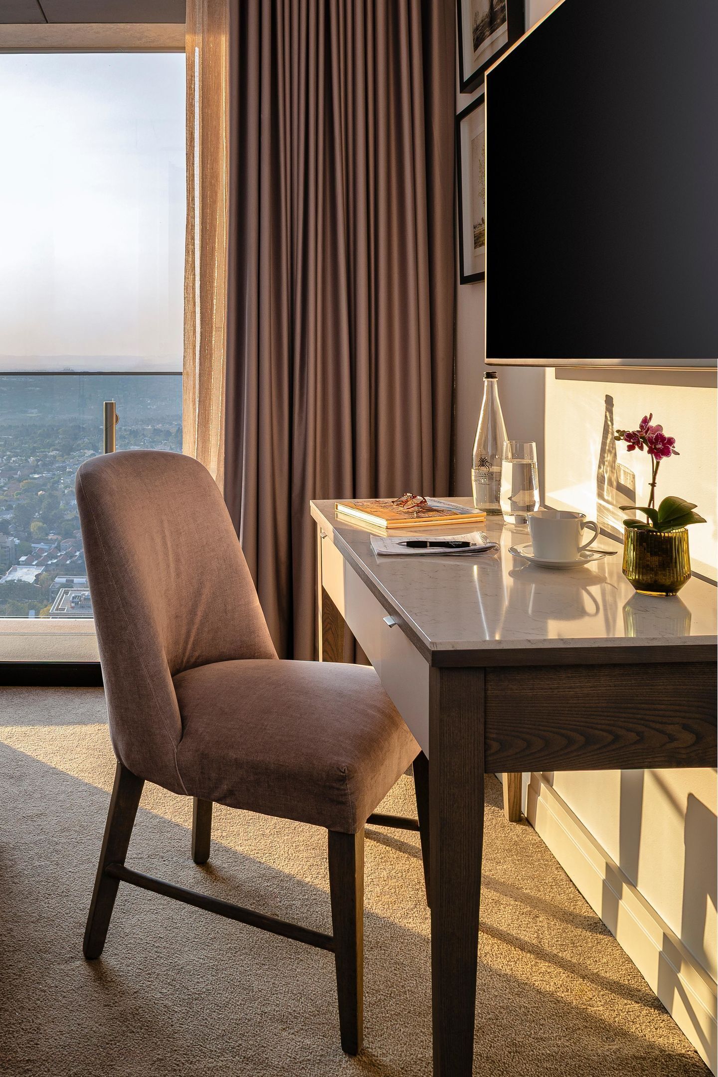 Una habitación de hotel moderna con un amplio ventanal que ofrece vistas a la ciudad, un sillón cómodo, un escritorio con una computadora portátil, una taza de café y un florero con una flor.