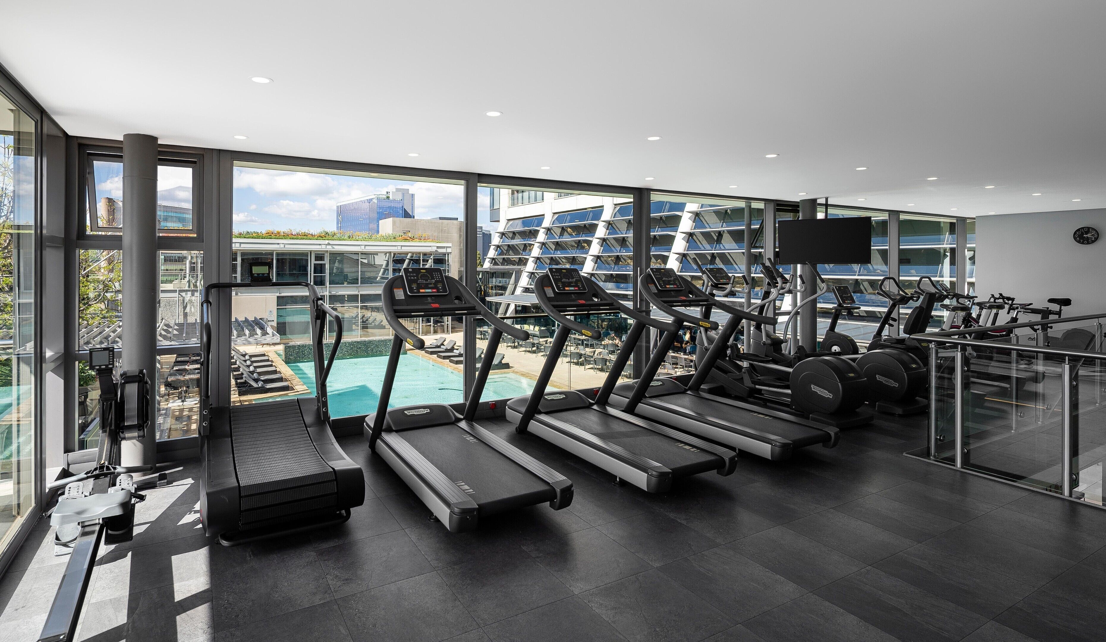 A modern gym with treadmills, exercise bikes, and a pool area visible through large windows.