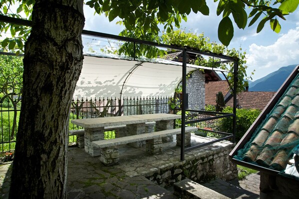 Un banc en pierre, placé sous une pergola surmontée d’une toile blanche, entouré d’un mur de pierre et d’une clôture en métal.