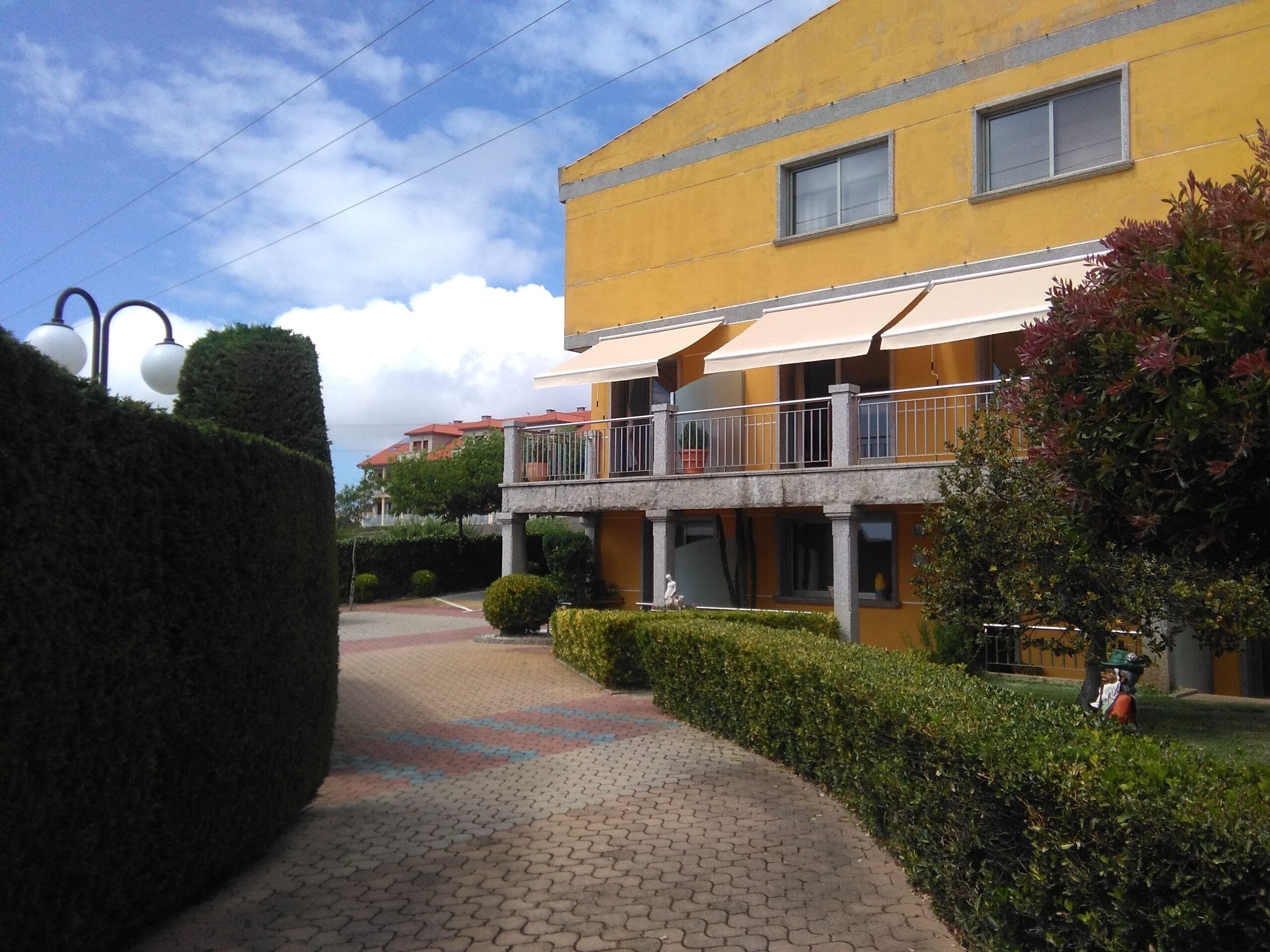 A yellow building with a balcony, a paved walkway, and neatly trimmed hedges.