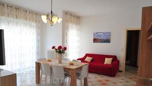 Een eetgedeelte met een houten tafel, witte stoelen en een vaas rode bloemen. Er is een rode bank en een schilderij aan de muur.