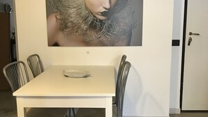 Un coin repas avec une table blanche et quatre chaises, un portrait encadré au mur et une porte sur la droite.