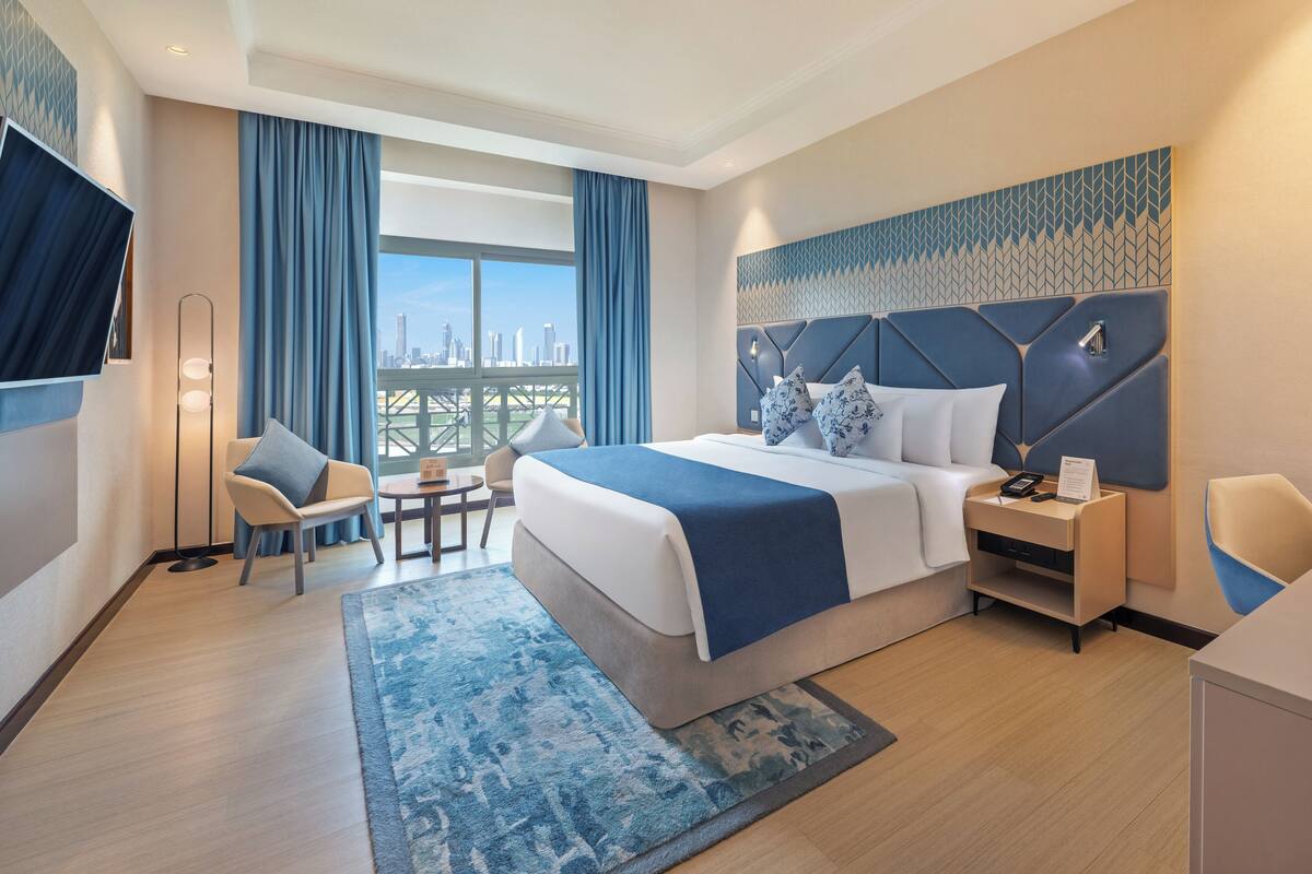 A modern hotel room with a large bed, a desk, a chair, a TV, and a view of the city skyline.
