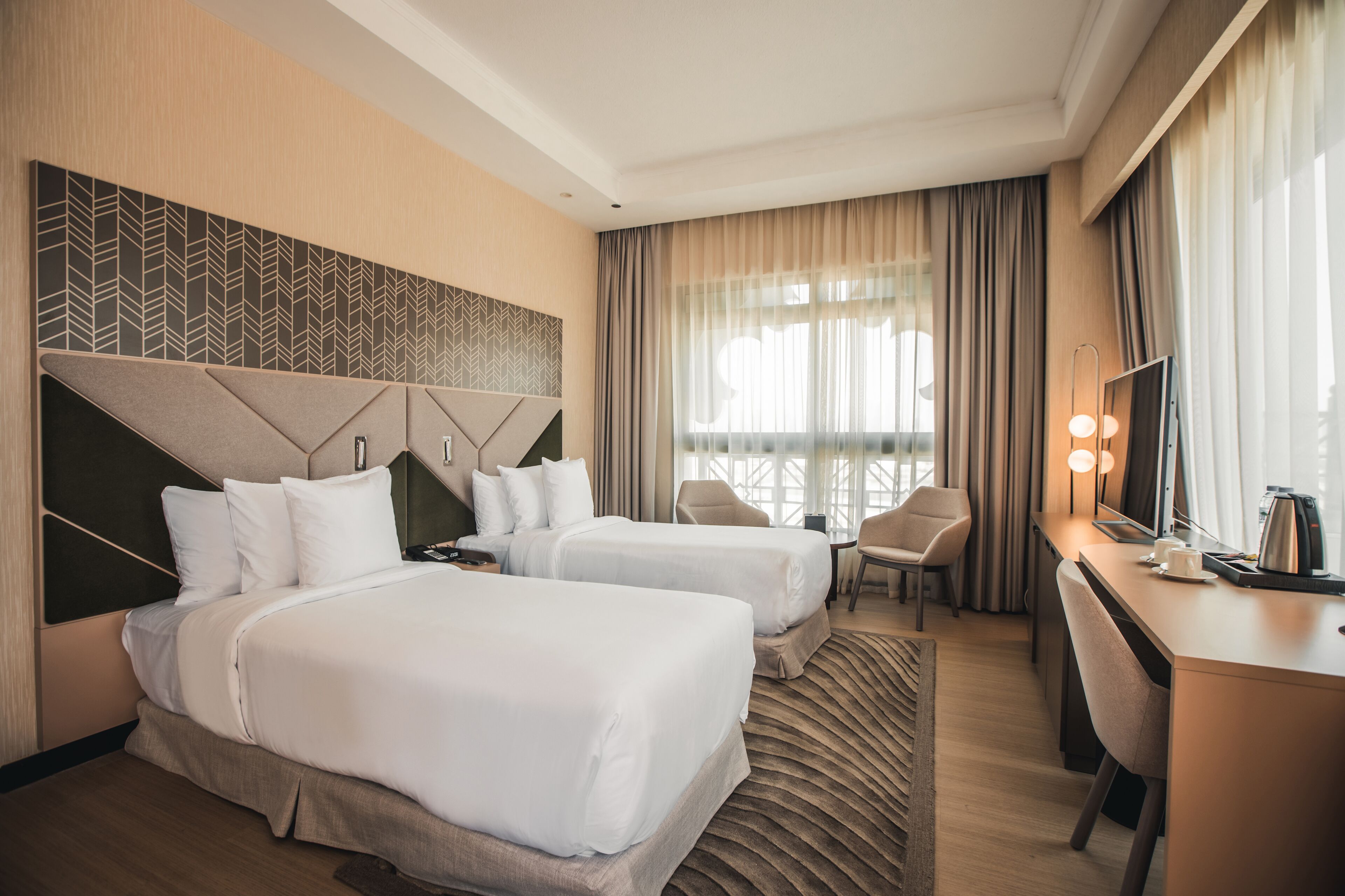 Habitación de hotel con dos camas, un escritorio, una silla y un ventanal con cortinas.