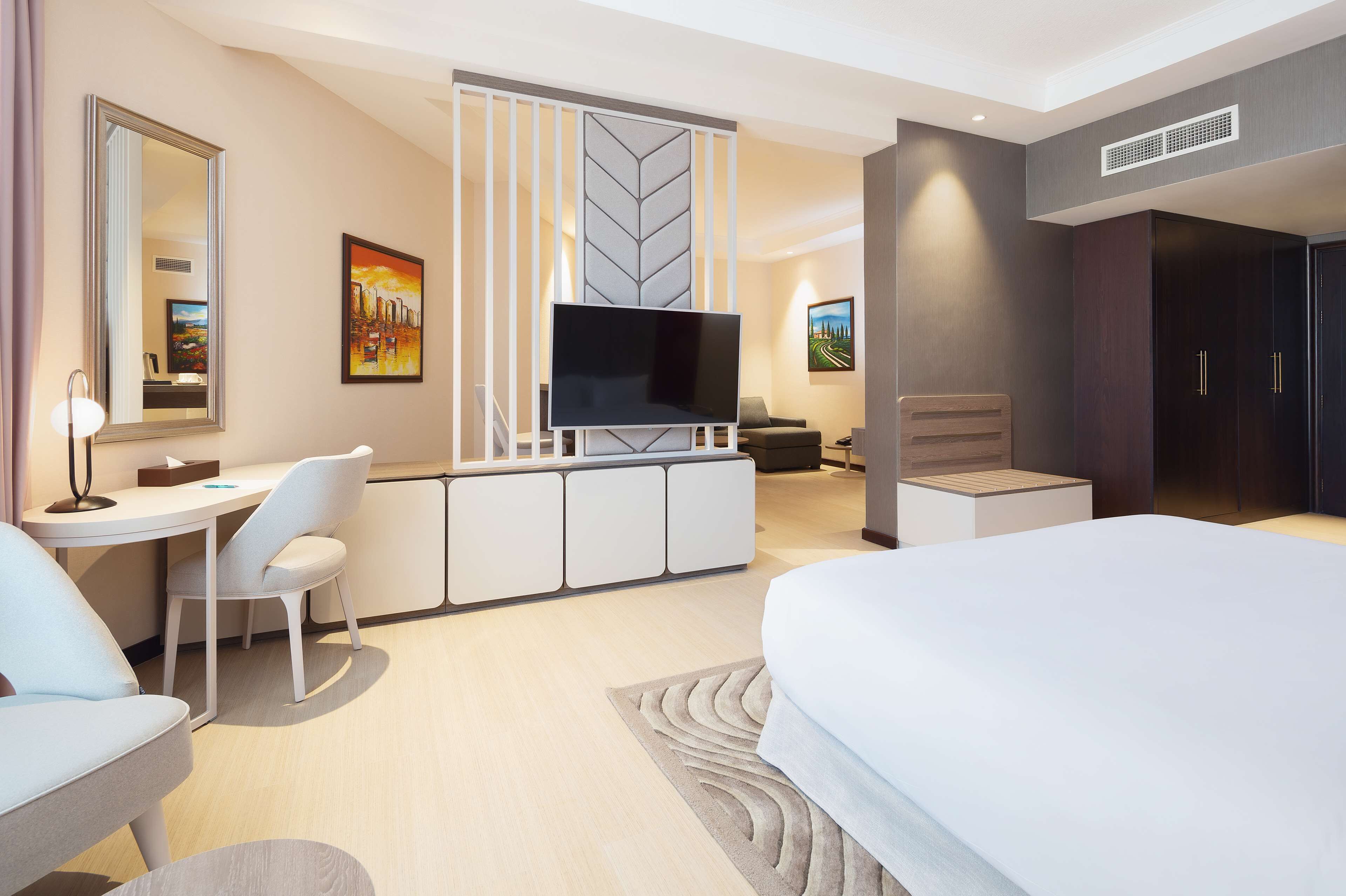 A modern hotel room with a large bed, a desk with a chair, a television, and artwork on the walls.