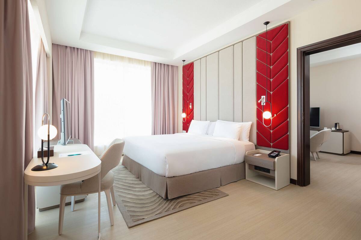 A modern hotel room with a neatly made bed, a desk with a lamp, a chair, and a small bedside table with a phone.