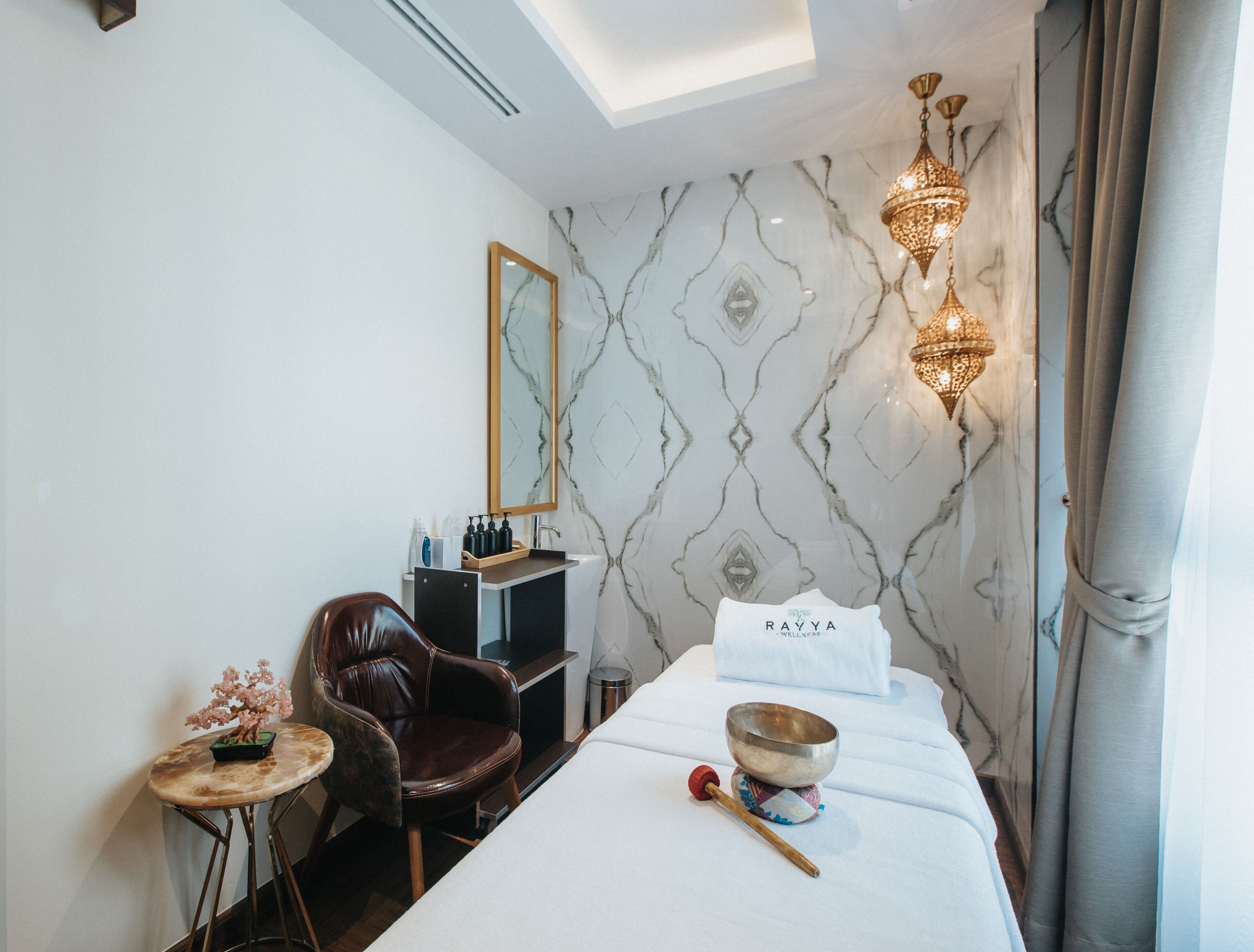 Une chambre d’hôtel moderne équipée d’un lit, d’une table de massage, d’un fauteuil de massage, d’une petite table avec un vase de fleurs, d’un miroir et d’un luminaire fixé au mur.
