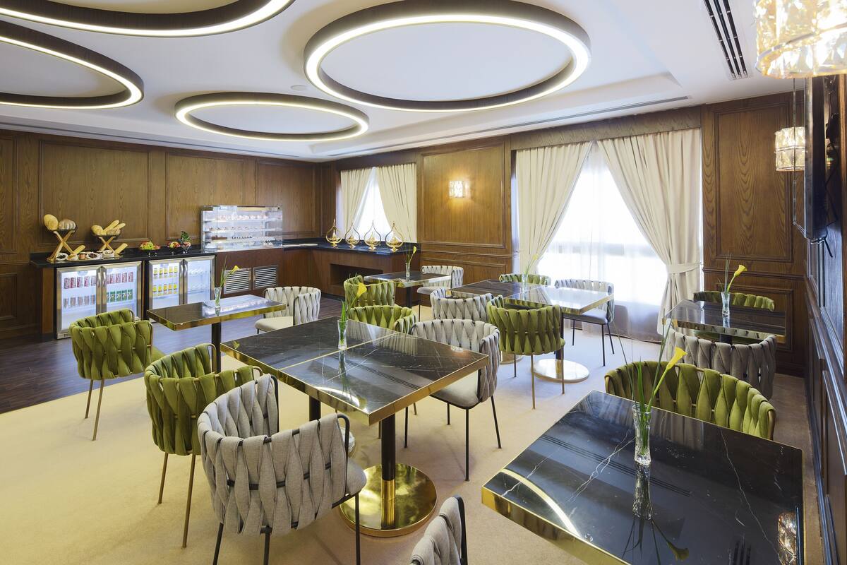 A modern hotel dining area with round tables and green upholstered chairs, a dessert display, and a view of the city through large windows.