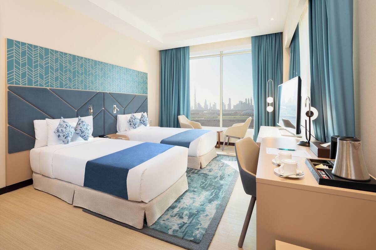 A hotel room with two beds, a desk with a coffee maker, a chair, and a large window with a city view.