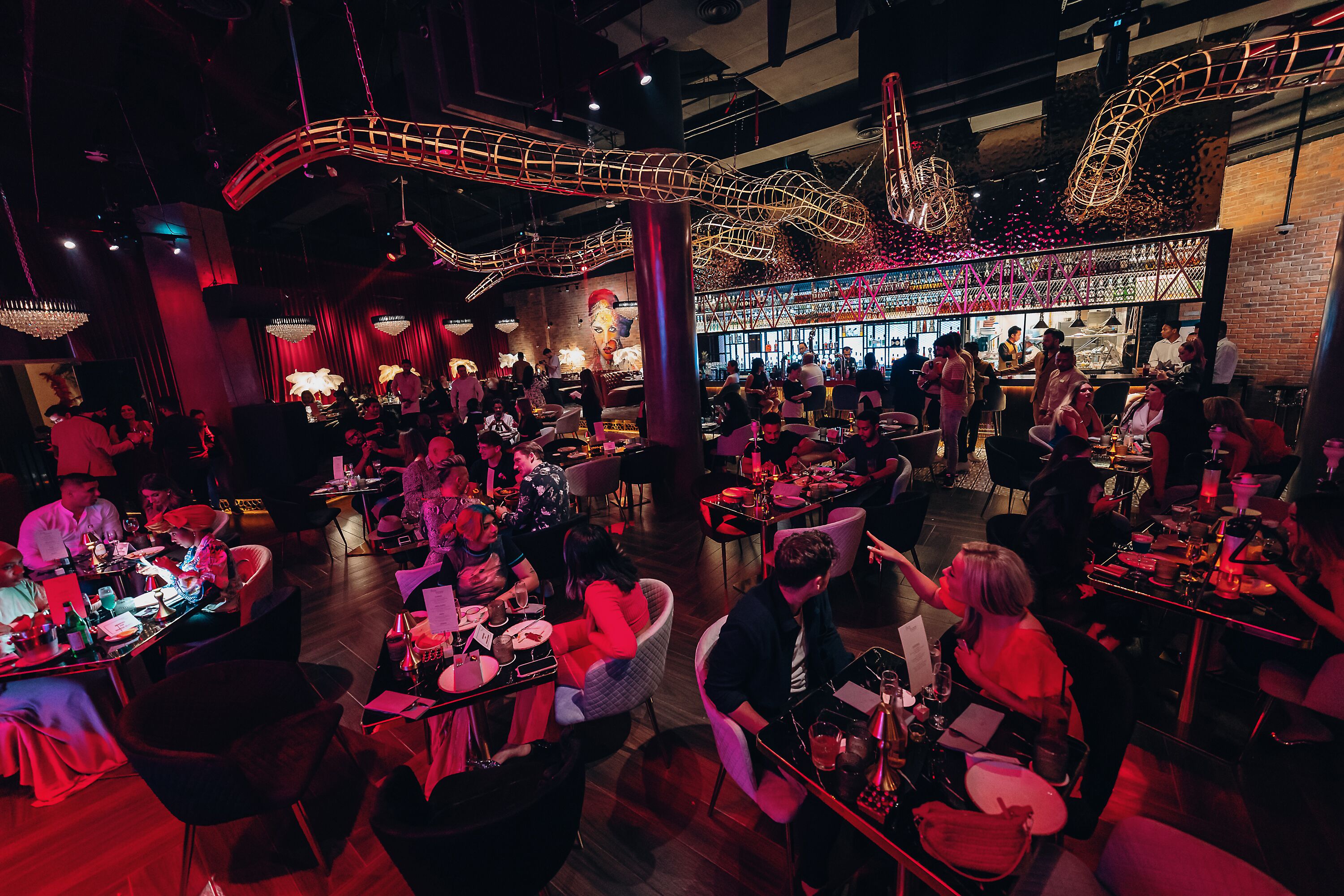 A bustling restaurant with multiple dining areas, a bar, and a large mural on the wall.