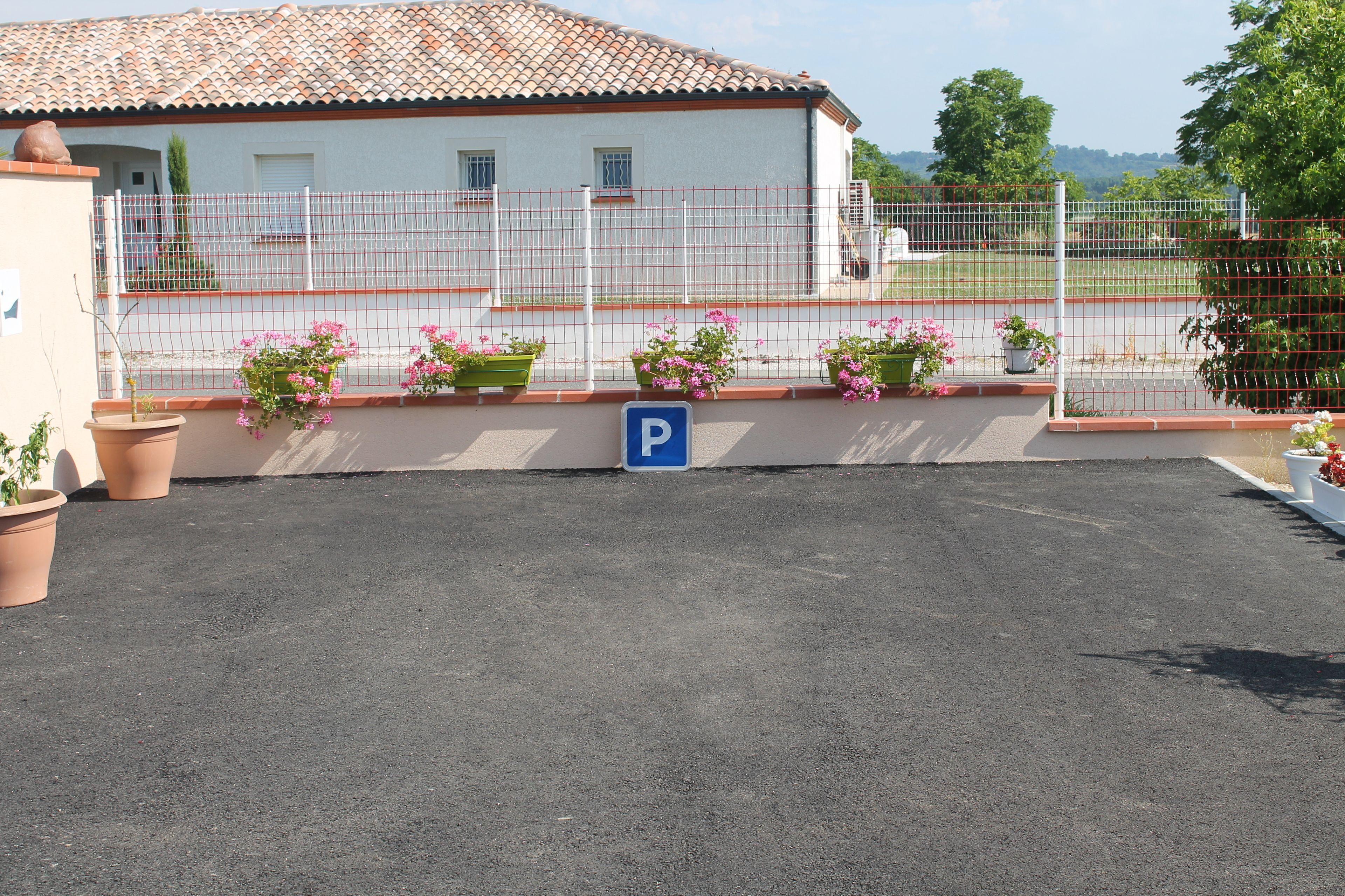 A parking area with a blue parking sign, potted plants, and a building with a tiled roof in the background.