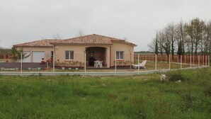 Uma casa térrea com telhado de telhas, varanda frontal com mobiliário de exterior e quintal cercado.