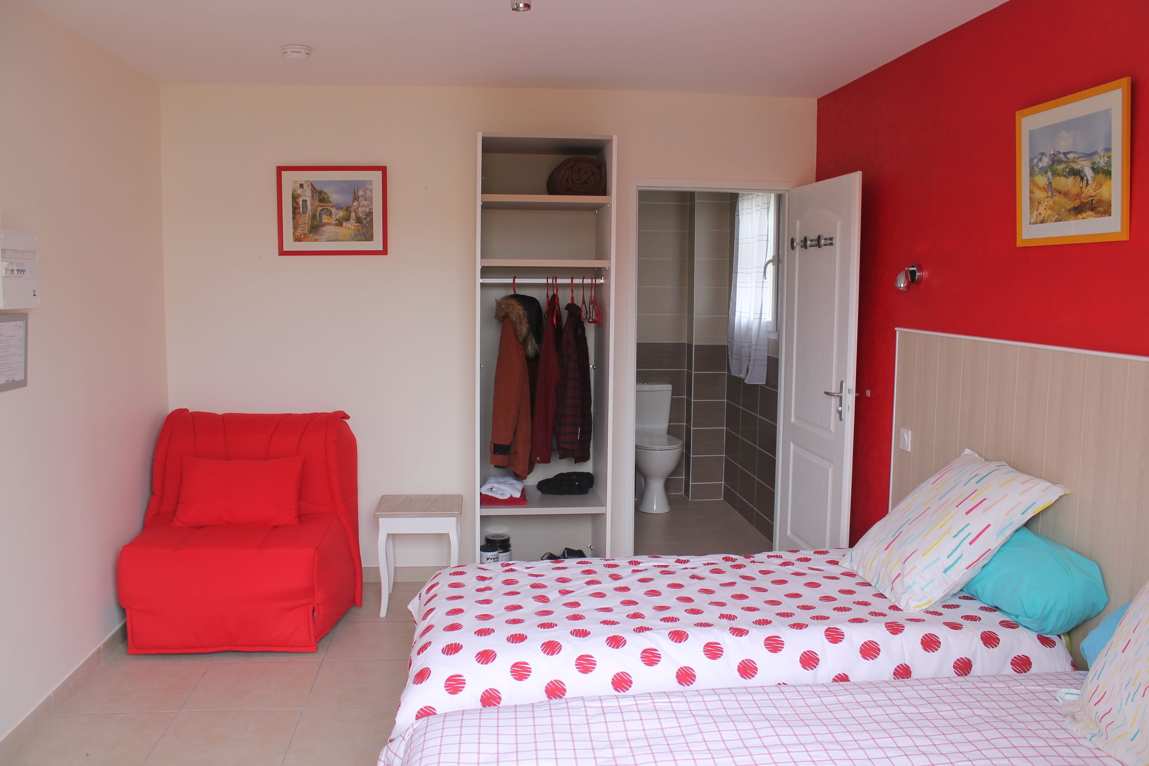 Un dormitorio con cama, una silla roja, un armario y un baño visible a través de una puerta abierta.