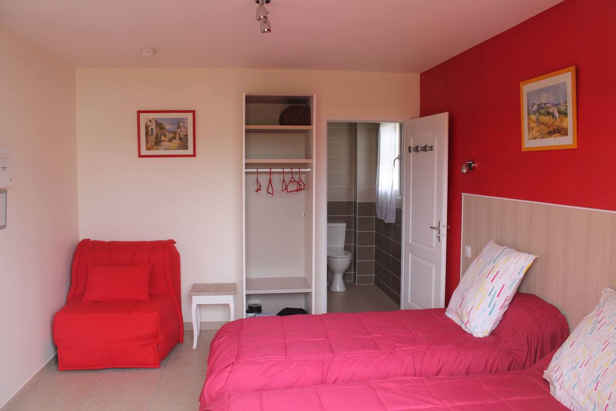 Une chambre à coucher avec un lit, une chaise rouge, une armoire et une salle de bain visible à travers une porte ouverte.