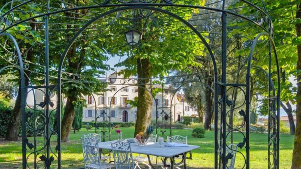 Un jardin aménagé avec une table et des chaises placées sous une arche métallique, entouré de verdure et avec un bâtiment en arrière-plan.