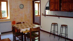 A dining area with a table and chairs, a clock on the wall, and a kitchen with wooden cabinets and stools. - The Temple at Solebello (Morro D' Alba)