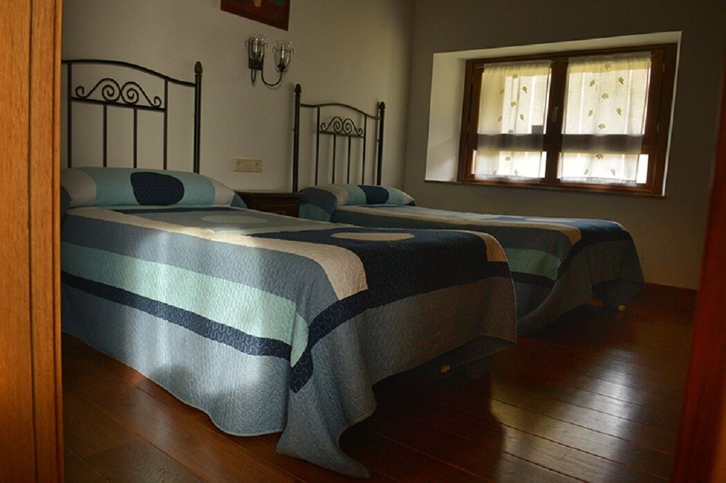 A room with two beds, wooden flooring, a window with curtains, and wall-mounted lighting.