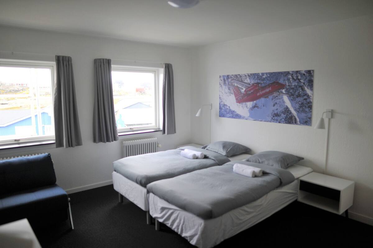 Ein Hotelzimmer mit zwei Betten, einem Fenster mit Vorhängen, einem Heizkörper, einem Schreibtisch und einem Bild an der Wand.