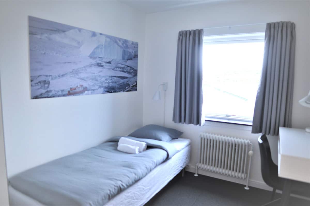 Ein kleines Zimmer mit einem Einzelbett, einem Fenster, einer Heizung und einem Schreibtisch mit Stuhl.