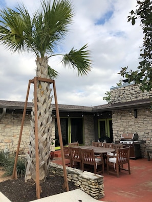 A patio area with a stone wall, a wooden table with chairs, a grill, and a palm tree.