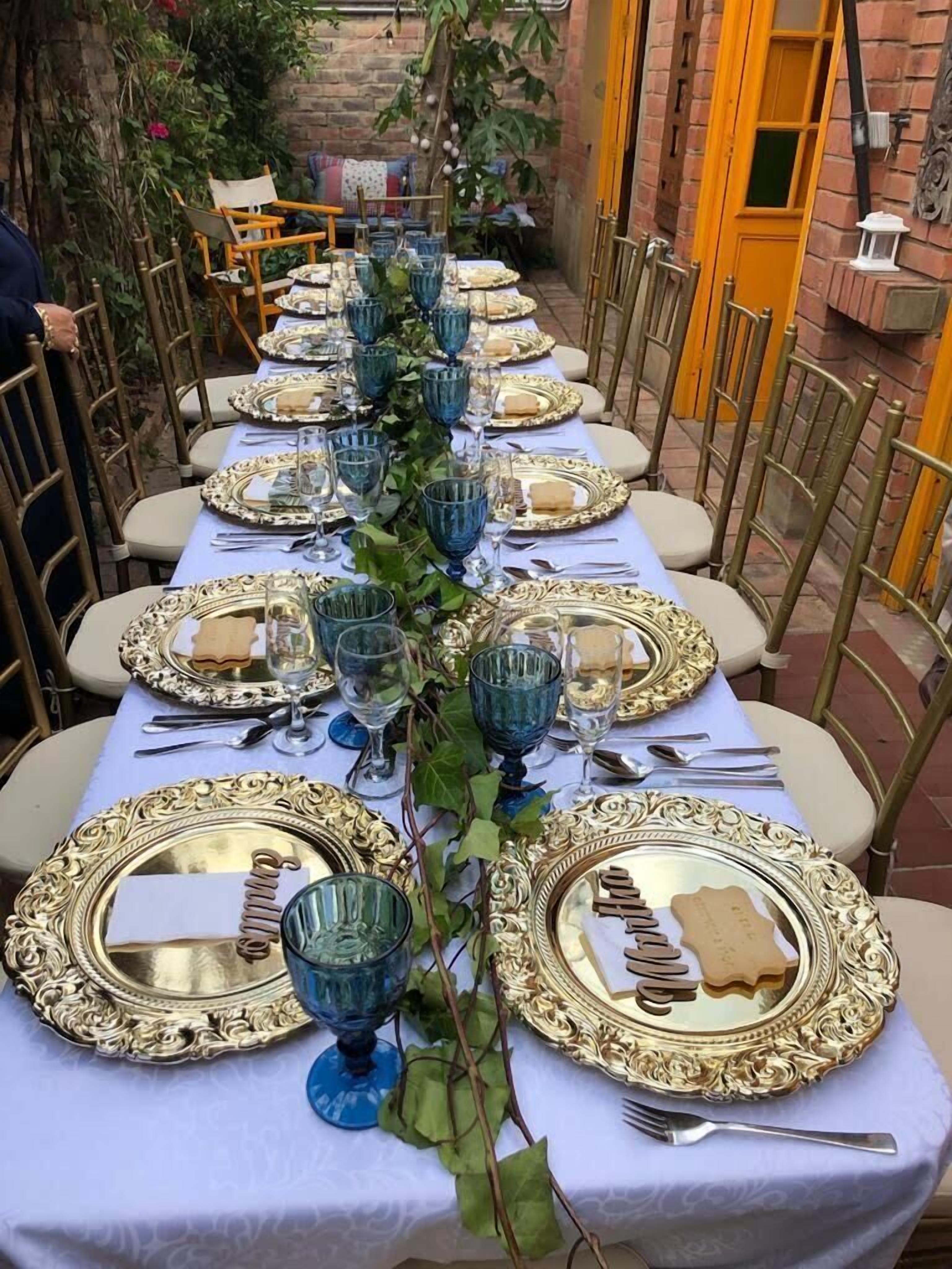 Mesa dispuesta para una cena al aire libre elegante, con platos de oro ornamentados y cristalería azul, rodeada de sillas, un muro de ladrillo y follaje al fondo.