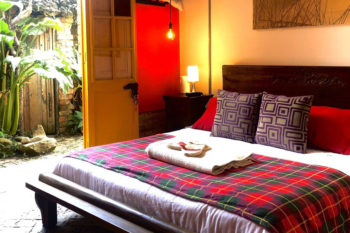 A bedroom with a bed, a nightstand, a lamp, a painting, and a view of the outdoors.