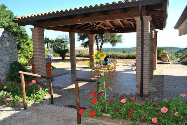 A covered outdoor seating area with brick pillars, surrounded by flowers and a view of the countryside.