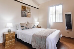 A bedroom with a bed, two bedside tables with lamps, a wooden dresser, a painting on the wall, a window with bars, and a door.