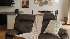 Ein modernes Wohnzimmer mit einem dunkelbraunen Ledersofa, einer beigen Decke und einem Makrameekissen. Im Hintergrund steht ein Esstisch mit Stühlen, und links ist ein Küchenbereich zu sehen.