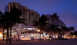A multi-story hotel with a modern design, featuring a prominent entrance with a fountain, surrounded by palm trees and other buildings.