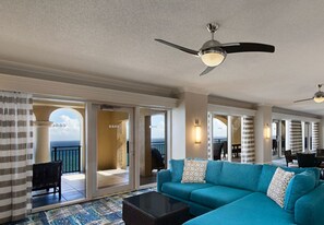 A spacious living area with a large blue sofa, a coffee table, and a ceiling fan. There is a view of the ocean through large glass doors.