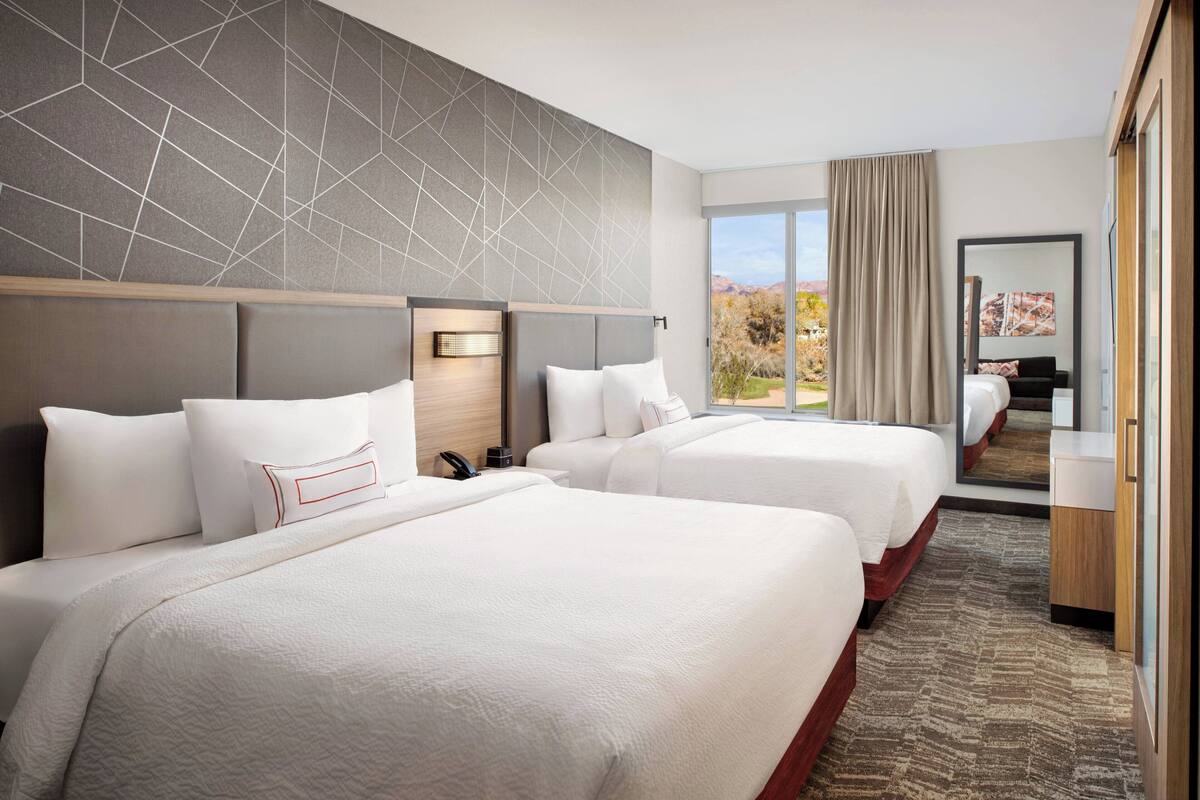 A hotel room with two beds, a large window with a view, a modern wall design, and a mirror reflecting the room.