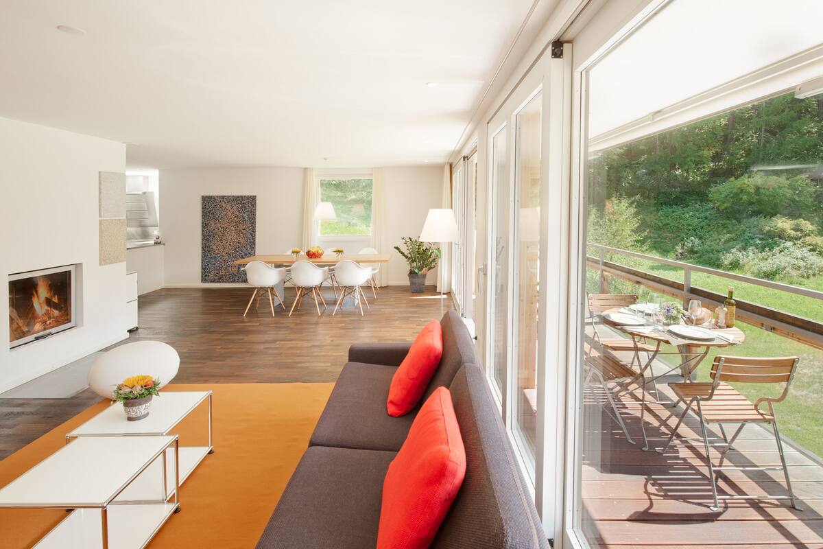 Ein modernes Wohnzimmer mit einer Couch, einem Esstisch und Stühlen, einem Kamin und einem Balkon mit Blick auf Grünflächen.