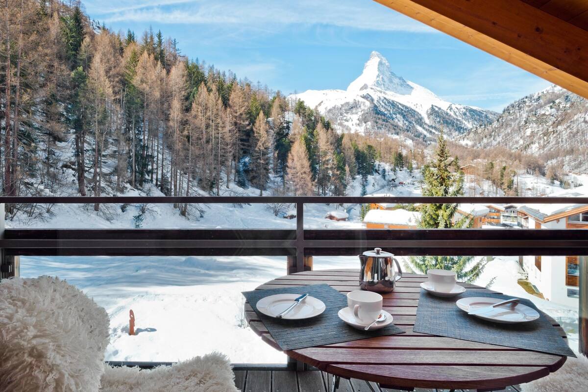 Eine Holzterrasse mit Tisch für Zwei, mit Blick auf eine verschneite Berglandschaft und einen markanten Berggipfel.