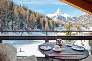 Een houten terras met een tafel gedekt voor twee, met uitzicht op een besneeerd berglandschap met een opvallende bergtop.
