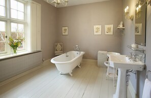 Ein Badezimmer mit freistehender Badewanne, Waschbecken und Heizkörper. An der Wand hängen Bilder in Rahmen, und ein Fenster bietet Blick ins Freie.