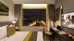 A hotel room with a bed, a desk, a chair, a TV, and a window with a city view. - Citadines Blue Cove Danang (Da Nang)